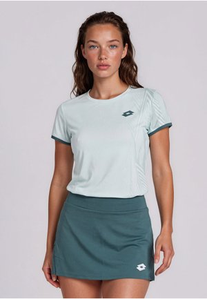 Jeune femme aux cheveux ondulés portant un t-shirt de sport vert clair à manches courtes et une jupe vert foncé, debout devant un fond uni.