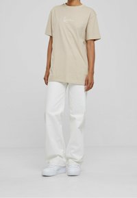 Camiseta de algodón beige con mangas cortas y diseño de logo blanco, combinada con pantalones anchos blancos y zapatillas blancas. Textura suave en general.