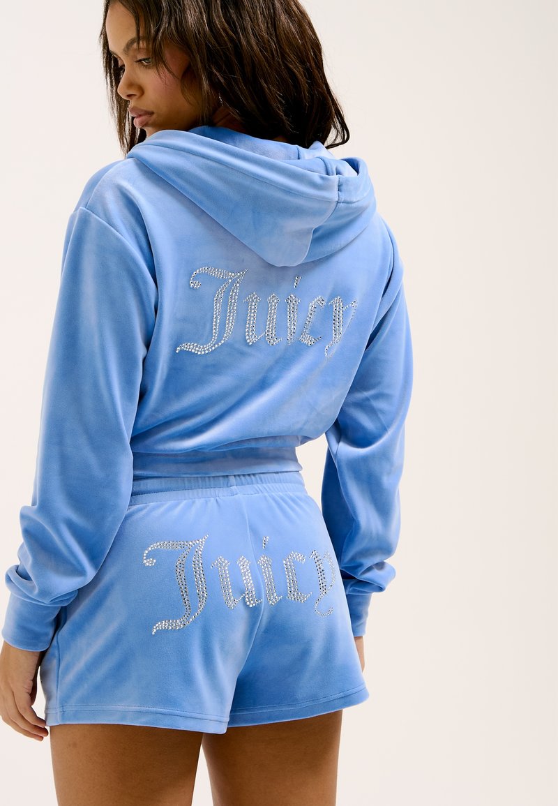 Tuta in velluto blu chiaro con cappuccio. Presenta la scritta "Juicy" in strass sul retro della parte superiore e dei pantaloni. Tessuto morbido, vestibilità comoda.