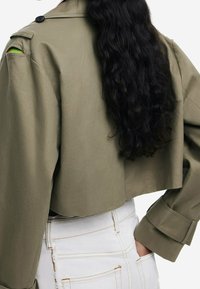 Chaqueta verde oliva recortada con estilo militar, hecha de tejido liso, que presenta un gran cuello, hombros estructurados y puños enrollados.