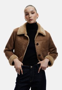 Brun ruskindsjakke med en cremegul pelskrave, to forlommer og to mørke knapper; parret med en sort turtleneck og denimjeans.