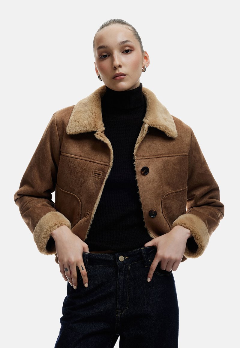 Brun ruskindsjakke med en cremegul pelskrave, to forlommer og to mørke knapper; parret med en sort turtleneck og denimjeans.