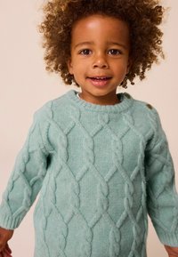 Pull en tricot bleu clair avec un motif en relief, col rond et couture visible. Comprend deux accents de bouton sur l'épaule.