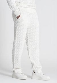Witte gebreide joggers met een gestructureerd kabelpatroon, voorzien van zijzakken en een geribde tailleband, gecombineerd met witte sneakers.