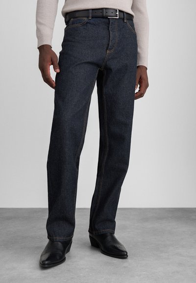 Jean en denim bleu foncé avec une coupe droite, couture visible et cinq poches, porté avec une ceinture noire et des bottes noires.