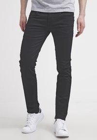 Černé slim-fit džíny vyrobené z denimu. Mají přední kapsy a zapínání na knoflík. Nosené s bílými teniskami na neutrálním pozadí.