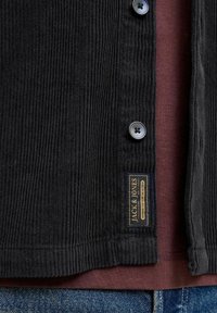 Chaqueta de pana negra con dos botones, que presenta una superficie texturizada y una etiqueta dorada. Llevada sobre una camiseta burdeos, combinada con jeans azules.