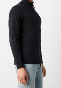 Schwarzer Reißverschluss-Pullover mit hohem Kragen, slim fit, gerippte Bündchen und Saum; getragen über hellblauen Jeans, aus weich strukturiertem Material.