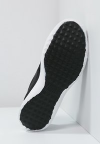 Svarta sneakers med texturerad gummisula, vit accent runt sulkanten och subtil märkning på sidan. Skorna lutade mot en vägg.