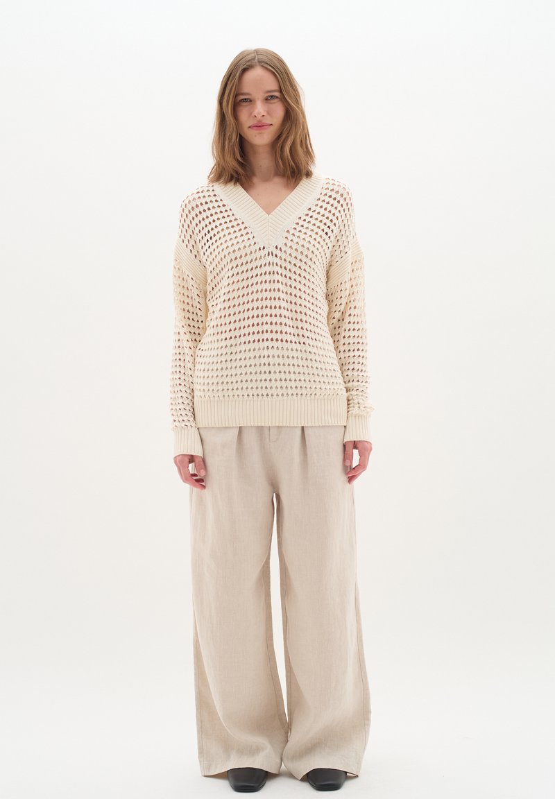 Strickpullover in Creme mit V-Ausschnitt, offenem Netzmuster und gerippten Bündchen, kombiniert mit beigen Schlaghosen und schwarzen Schuhen.