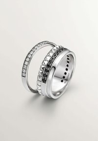 Tres anillos apilados: una banda de plata con tachuelas negras, una banda adornada con cristales y una banda de plata lisa. Presenta un acabado pulido y texturas variadas.