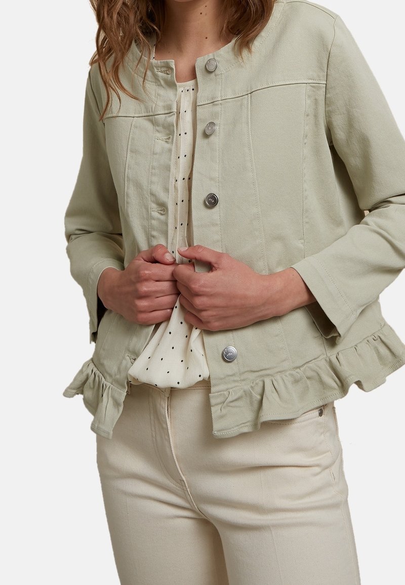 Femme portant une veste vert clair boutonnée avec un ourlet à volants sur un chemisier blanc à pois, associée à un pantalon crème, les mains tenant les bords de la veste.