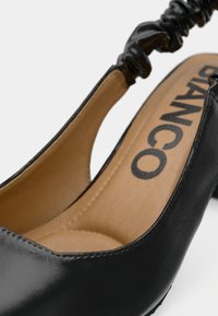 Μαύρο τακούνι από λουστρίνι με ανοικτή πλάτη και μαλακή μπεζ επένδυση. Διαθέτει λουράκι με πιεσμένες λεπτομέρειες για επιπλέον υφή. Λογότυπο στο εσωτερικό πέλμα.