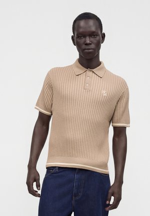 Mand iført en beige kortærmet strikket polo og mørkeblå jeans står mod en ensfarvet lys baggrund.