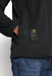 Černá bunda Adidas s texturovanou látkou, se zipovými kapsami a zlatým logem na boku. Nositá přes modré džíny.