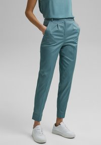 Pantalons aqua en tissu lisse, avec des poches latérales, des plis à l'avant et une coupe fuselée, portés avec des baskets blanches.