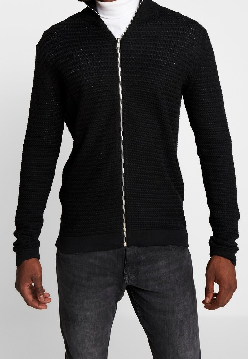 Man som bär en svart texturerad zip-up tröja över en vit polo, i kombination med mörkgrå jeans, visad från nacke till höfter.