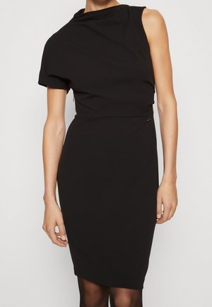 Woman wearing a fitted black sleeveless dress with an asymmetrical neckline and one short sleeve, standing against a plain background.