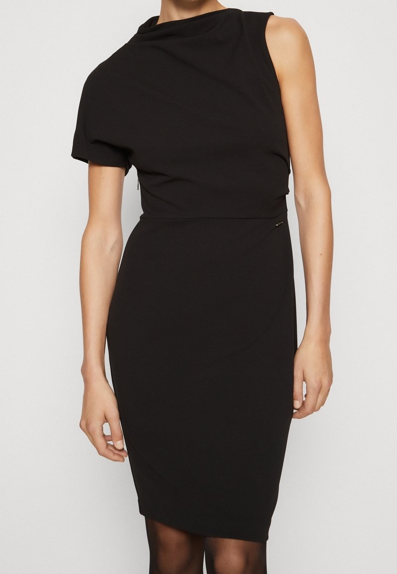 Femme portant une robe noire ajustée sans manches avec un décolleté asymétrique et une manche courte, debout devant un fond uni.