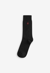 Black sock with a ribbed top, featuring a small red emblem. Made from a smooth material, with a classic, fitted shape and no patterns.