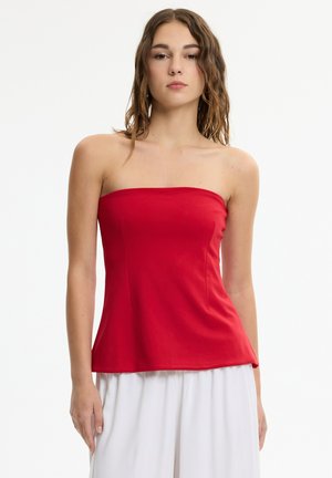 Mujer con cabello ondulado hasta los hombros, lleva un top rojo sin tirantes y pantalones blancos sueltos sobre un fondo blanco liso.