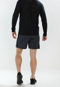 Black long-sleeve shirt paired with gray athletic shorts, featuring a relaxed fit. Model wears black sneakers. Background is plain white.