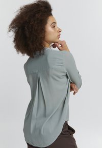 Blusa azzurro chiaro in tessuto morbido e fluido, con una piega sul retro e orlo arrotondato, maniche lunghe e un design minimalista.