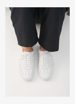 Tenisi din piele albă, cu o textură netedă, vârf rotund și un sistem de șireturi clasic, poziționați deasupra unor pantaloni oscuri, largi.