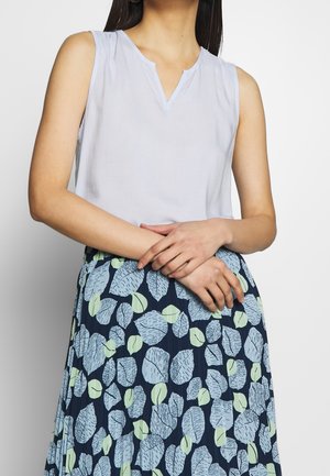 Žena nosící bílý nátělník bez rukávů a námořnickou sukni s velkými světle modrými a zelenými listovými vzory, ruce jemně překřížené v pase.