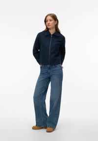 Femme portant une veste bleu marine zippée, un jean à jambes larges bleu et des chaussures marron clair, debout avec les mains derrière le dos devant un fond blanc uni.