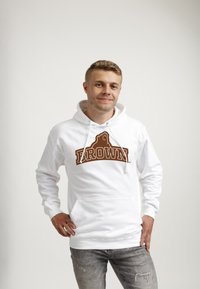 Biała bluza z kapturem z brązowym logo "BROWN" i grafiką niedźwiedzia, z kieszenią kangurka i sznurkiem w kapturze. Noszona z jasnoszarymi dżinsami.