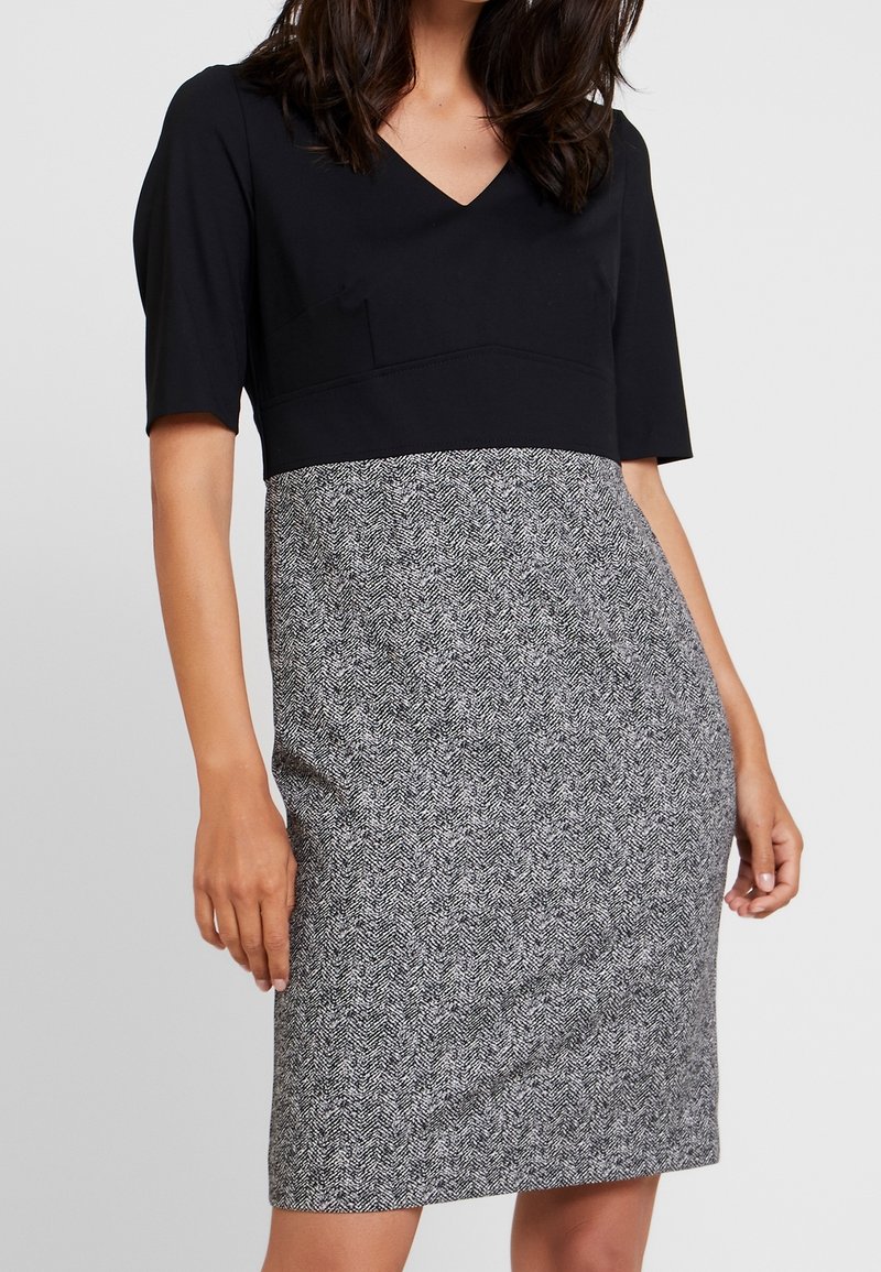 Femme portant une robe longueur genoux avec un haut noir ajusté et une jupe grise à motif chevron, debout devant un fond uni.