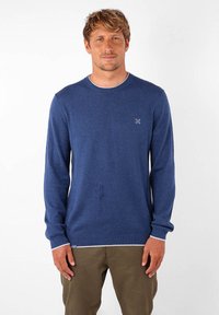 Pull en maille bleu marine à manches longues, col rond, avec des bordures blanches aux poignets et à l'ourlet. Présente un petit logo sur la poitrine.