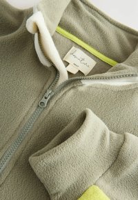 Fleece striukė švelniai žalia spalva, su minkštu baltu pamušalu, turi užtrauktuką ir ryškiai geltoną akcentą prie apykaklės. Viduje matomas etiketė.