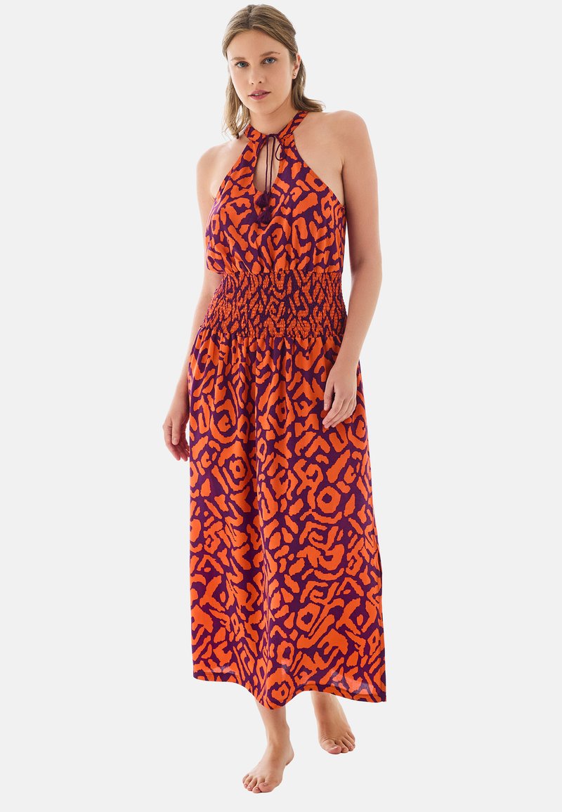 Mujer con vestido largo sin mangas de cuello halter con estampado abstracto en naranja y púrpura oscuro, de pie descalza sobre un fondo liso.