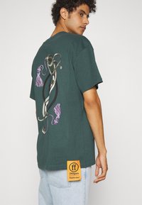 Musta vihreä lyhythihainen t-paita, jossa on graafinen design takana, oranssi label helman kohdalla, ja rento leikkaus.