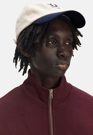 La gorra tiene una parte delantera en color crema y un visera en azul marino, hecha de algodón. El modelo lleva una chaqueta de cremallera color marrón, mostrando un perfil de cerca.