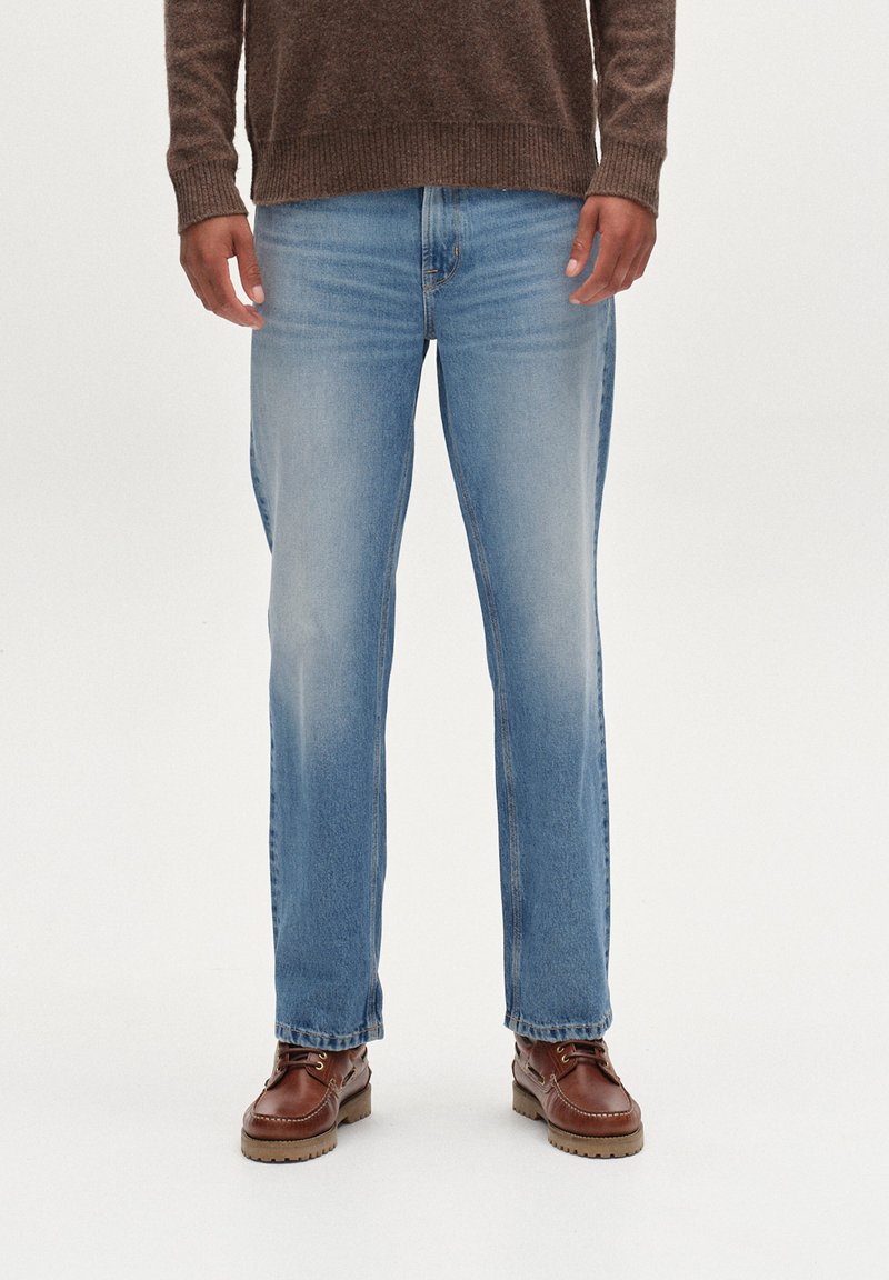 Jeans de mezclilla blue con corte recto, que presentan un lavado claro y un ligero desgastado. Combinados con zapatos de piel marrón y un suéter marrón oscuro.