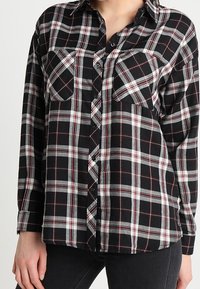 Black, white, and red plaid long-sleeve button-up shirt with two chest pockets worn with black pants.