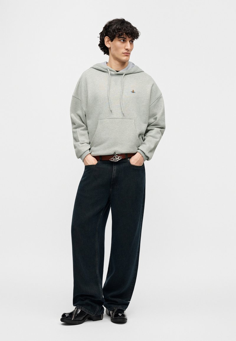 Sweat à capuche gris avec une poche avant, associé à un jean large bleu foncé et des chaussures noires. La ceinture remarquable présente une boucle décorative.