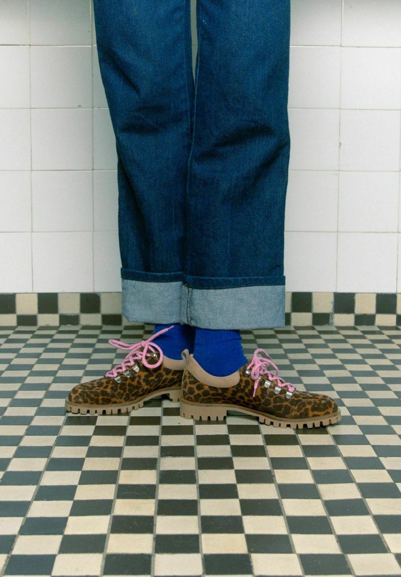 Scarpe con stampa leopardata, lacci rosa e dettagli beige, abbinate a calzini blu e jeans in denim scuri risvoltati su un pavimento a piastrelle bianche e nere.