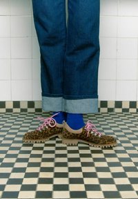 Scarpe con stampa leopardata, lacci rosa e dettagli grigi, abbinate a calzini blu, indossate con jeans in denim scuro con risvolti su un pavimento a scacchi.