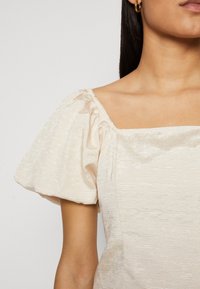 Top con maniche a sbuffo e scollatura quadrata in tessuto beige chiaro testurizzato con un leggero luccichio. Presenta maniche arricciate e un design della parte superiore aderente.