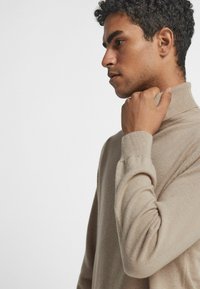 Beige höghalsad tröja i mjukt material, med åtsittande muddar och en subtil textur, visad på en modell i profil.