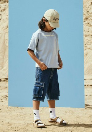 Laps, kes kannab beežikat mütsi, helerohelist T-särki, teksaste laadseid lühikesi pükse, valgeid sokke ja sandaale, seisab liivasel pinnal sinise tausta ees.