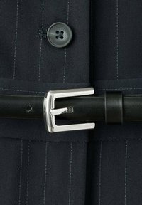 Gros plan sur une veste de costume sombre à fines rayures avec un bouton noir et une ceinture en cuir noire avec une boucle métallique argentée.