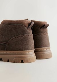 Brūni suede potītes zābaki ar teksturētu apdari, ar kontrastējošu brūna krāsas zoli, noapaļotu papēdi un diviem cilpu tabiem aizmugurē.