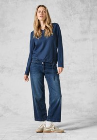 Marineblaues Langarmshirt aus strukturiertem Stoff mit gerafftem Taillenbund, kombiniert mit lockeren, dunkelblauen Jeans und hellbeigen Sneakers.