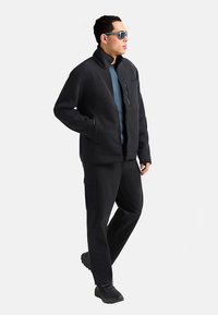 Giacca nera a coste con colletto alto, tasca con zip, abbinata a pantaloni scuri e calzature nere, indossata da una persona con occhiali da sole.