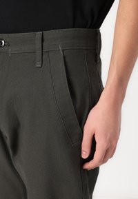 Ciemnozielone spodnie z mieszanki bawełny, wyposażone w gładki przód, boczne kieszenie oraz zapięcie na guzik. Gładka faktura z minimalnymi detalami szwów.